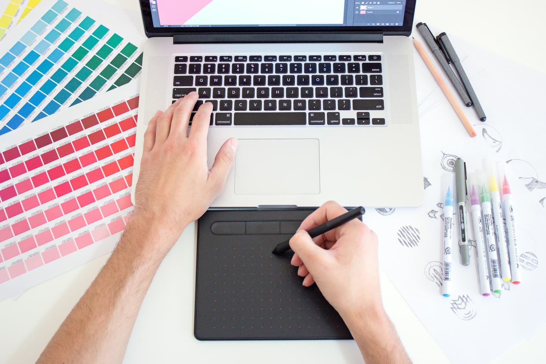 A designer working with a pen tablet beside colour palettes and a laptop.