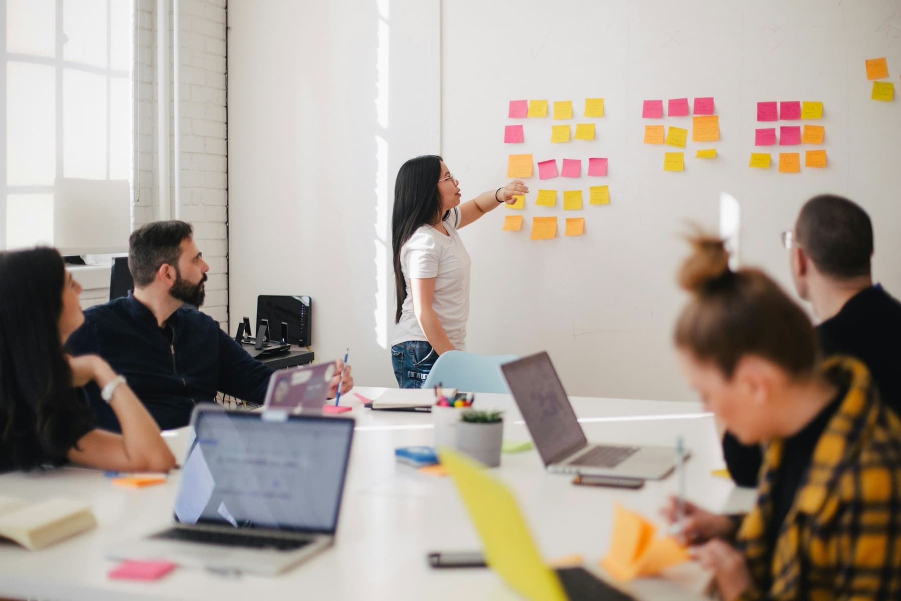 A team workshop: one person presents ideas on sticky notes while colleagues take notes at their laptops.